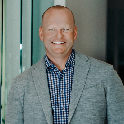 Man in gray blazer and plaid shirt smiles in a modern setting.