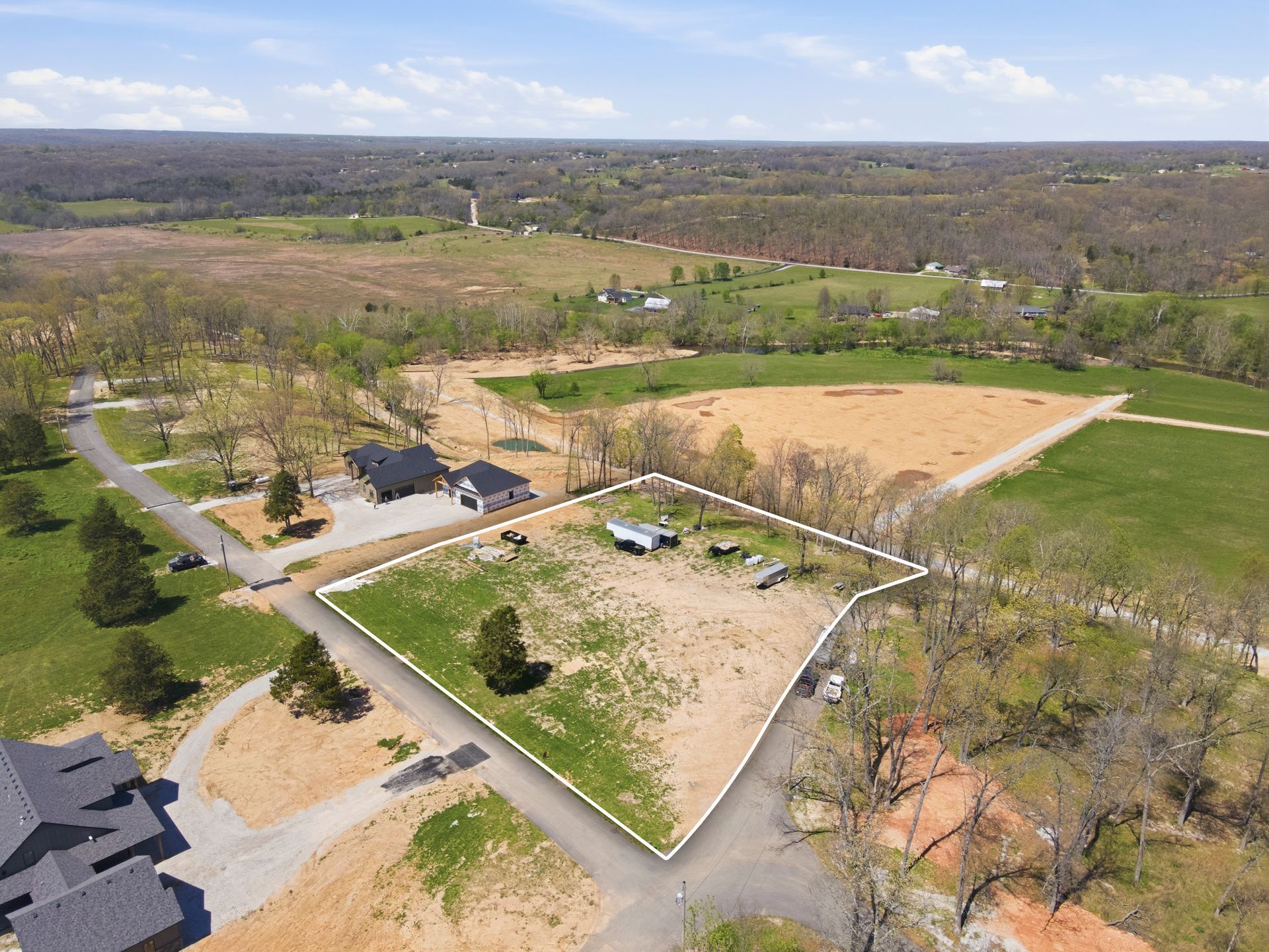 Aerial view of a rural property with a fenced field, gravel roads, trees, and a barn under a blue sky