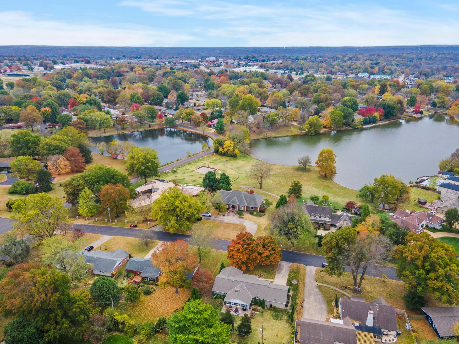 Aerial view of a suburban neighborhood with houses, colorful trees, and two connected lakes under a cloudy sky.