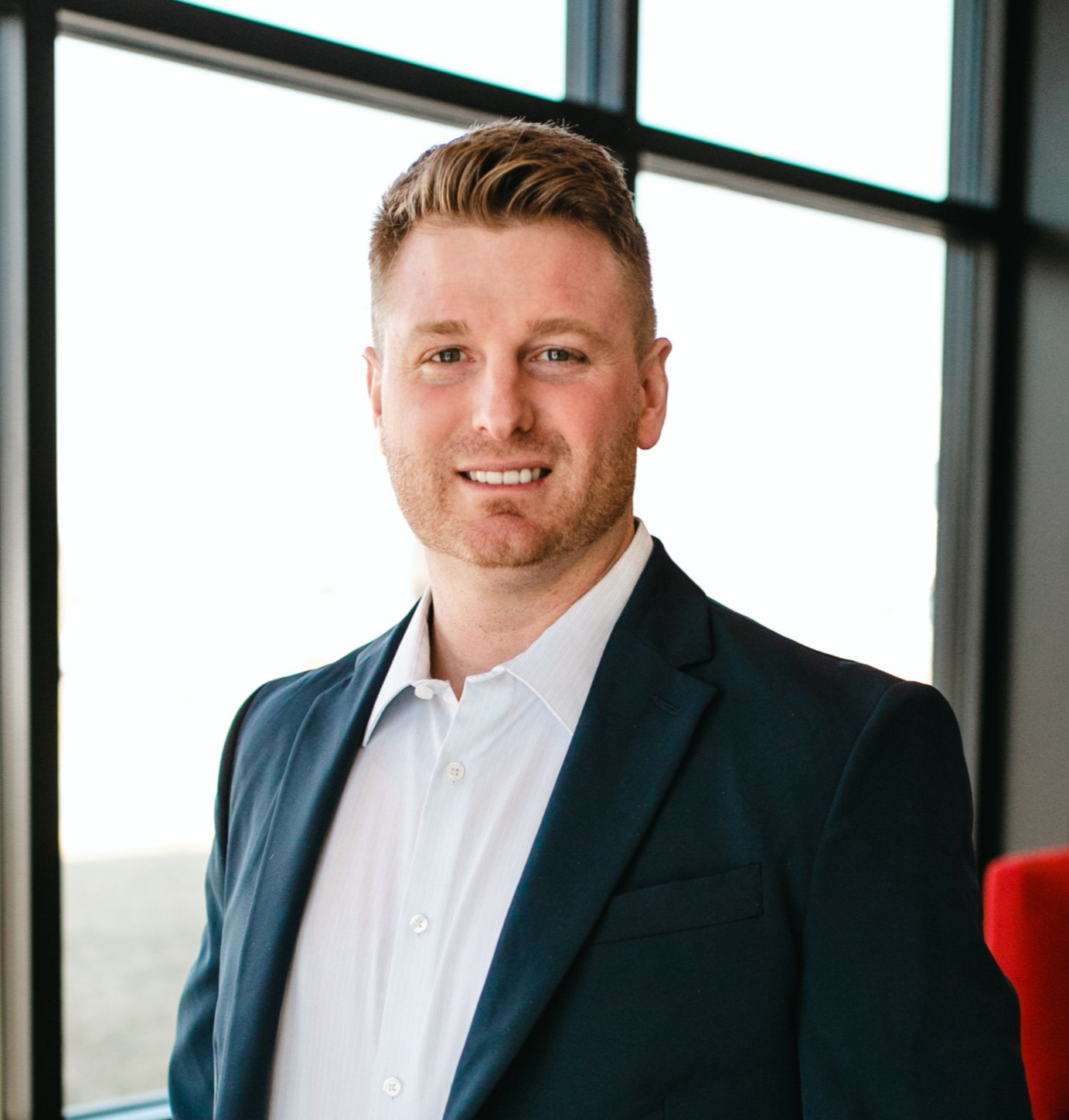 Man in gray blazer and plaid shirt smiles in a modern setting.