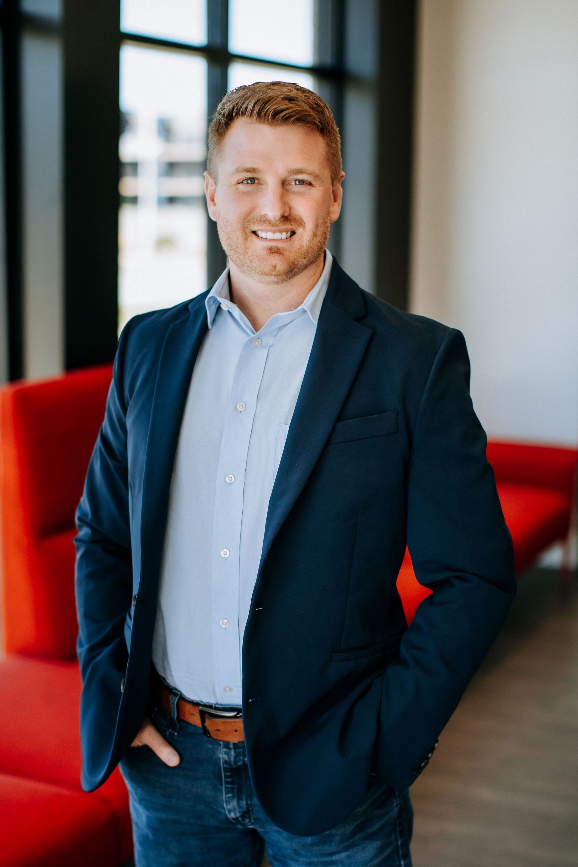 Man in a navy blazer and jeans, smiling, posing with hands in pockets; red chairs in the background.