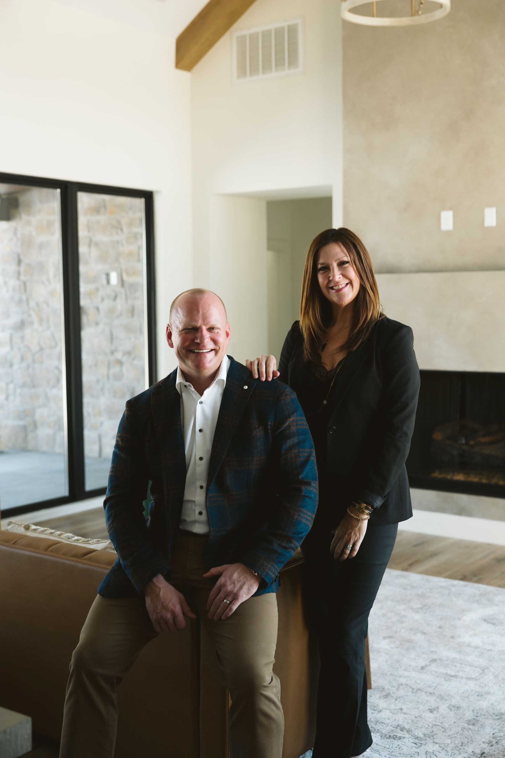 Man in blazer sits, woman stands nearby in a modern interior; they smile.