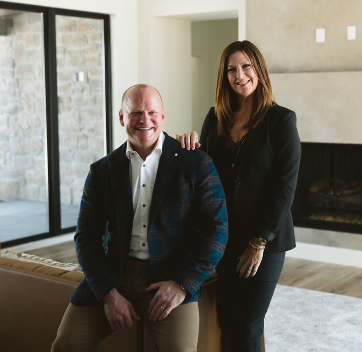 Two people posing in a modern living room, smiling by a large fireplace and glass doors