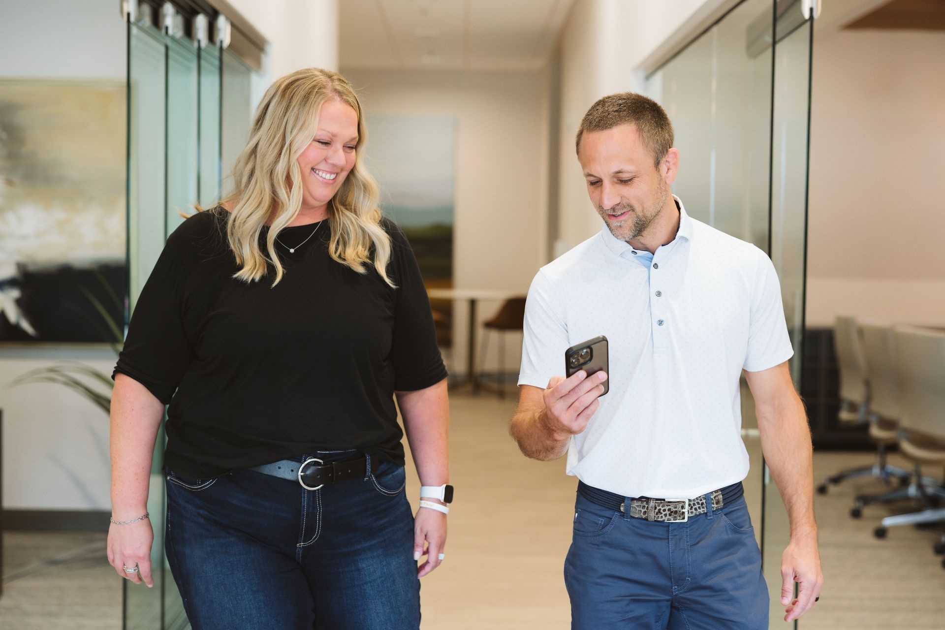 Woman and man walking in office hallway, man looking at phone, both smiling.