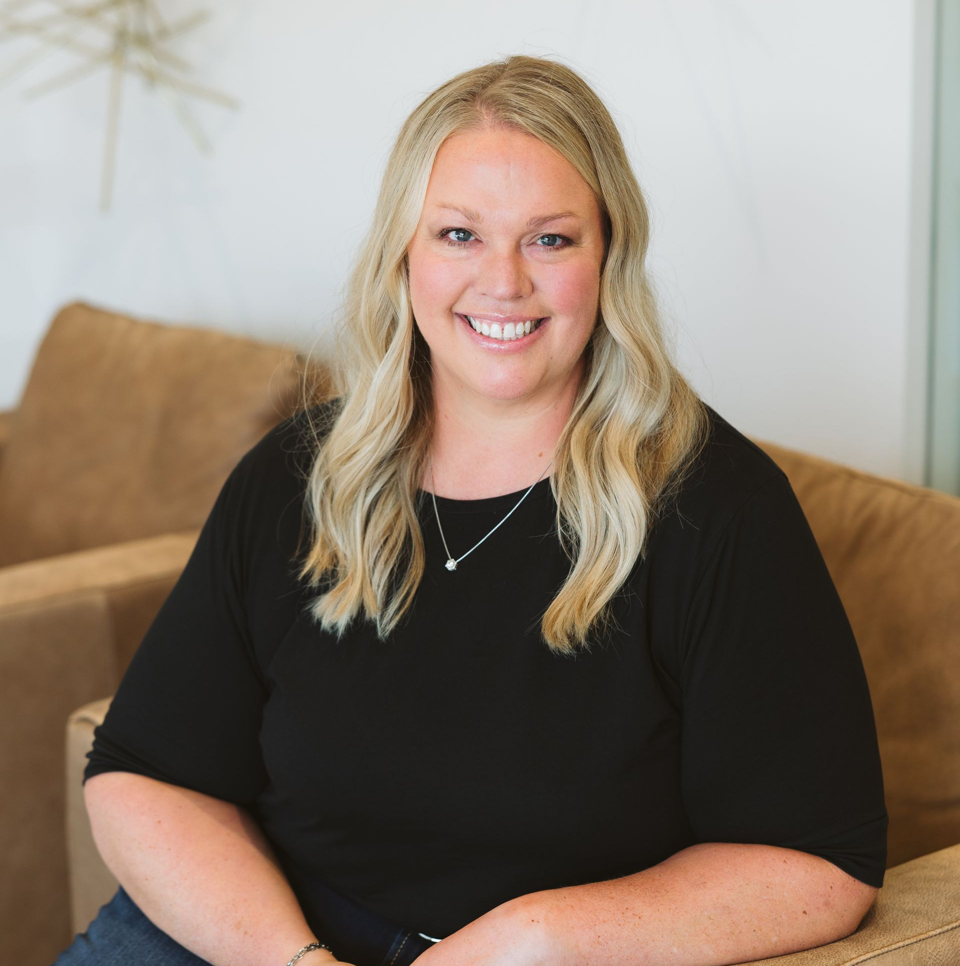 Woman with blonde hair smiling, wearing a black top, seated on a brown couch.