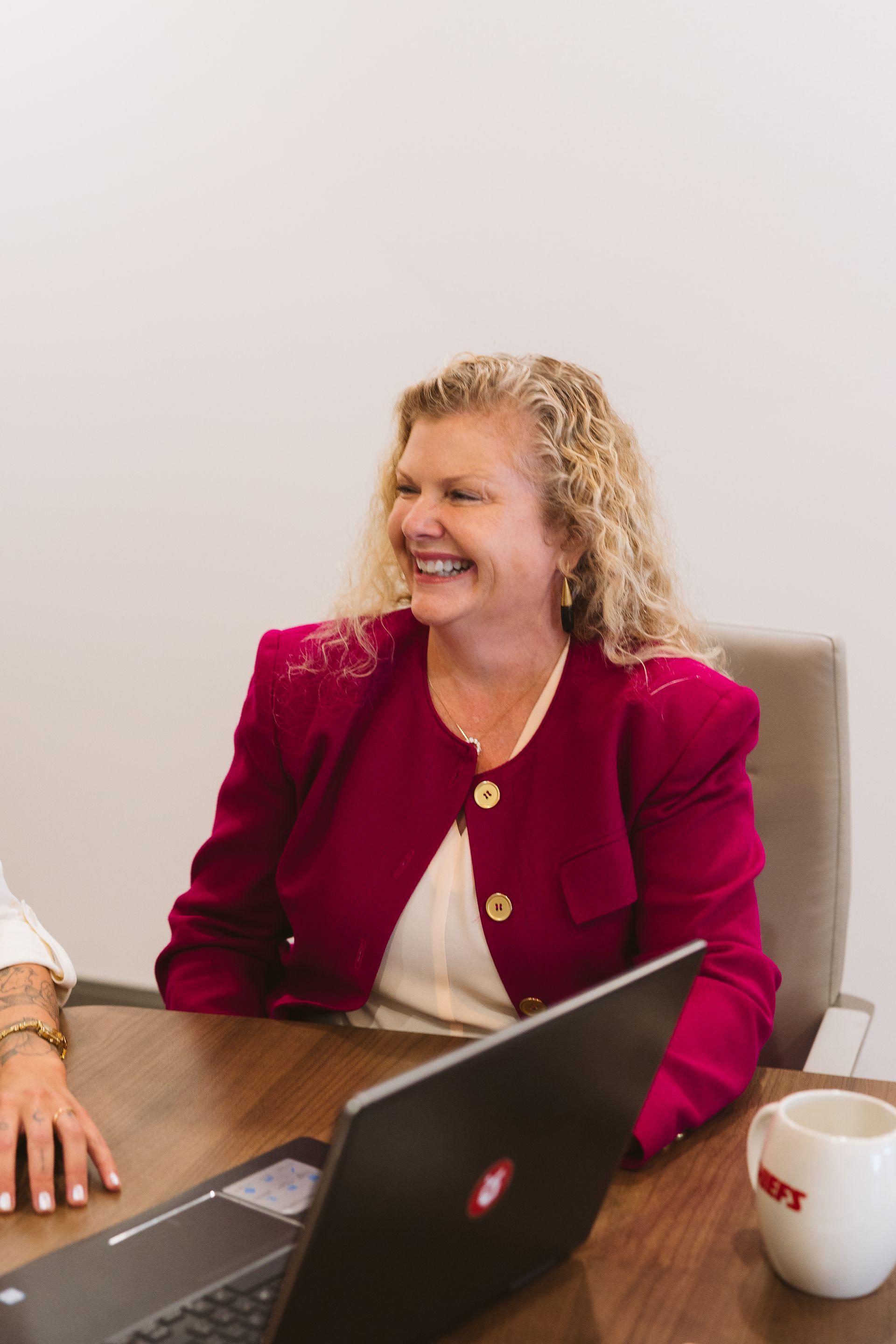 Woman with curly blonde hair smiles, wearing a black jacket and teal top, indoors near a red sofa.