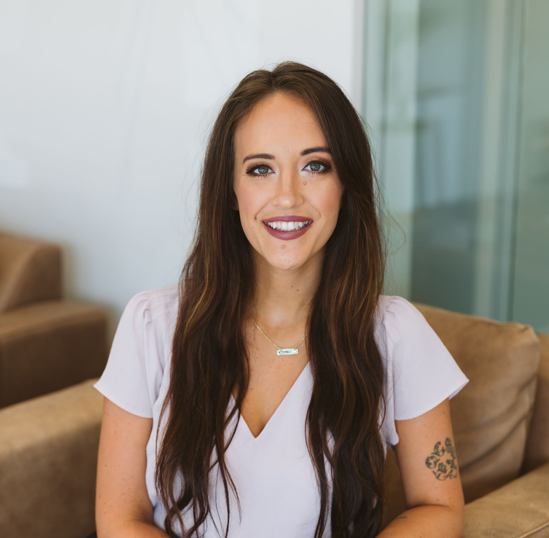 Woman with light brown hair smiles, wearing a black top and a necklace, in a bright setting.