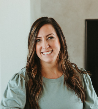 Woman with light brown hair smiles, wearing a black top and a necklace, in a bright setting.