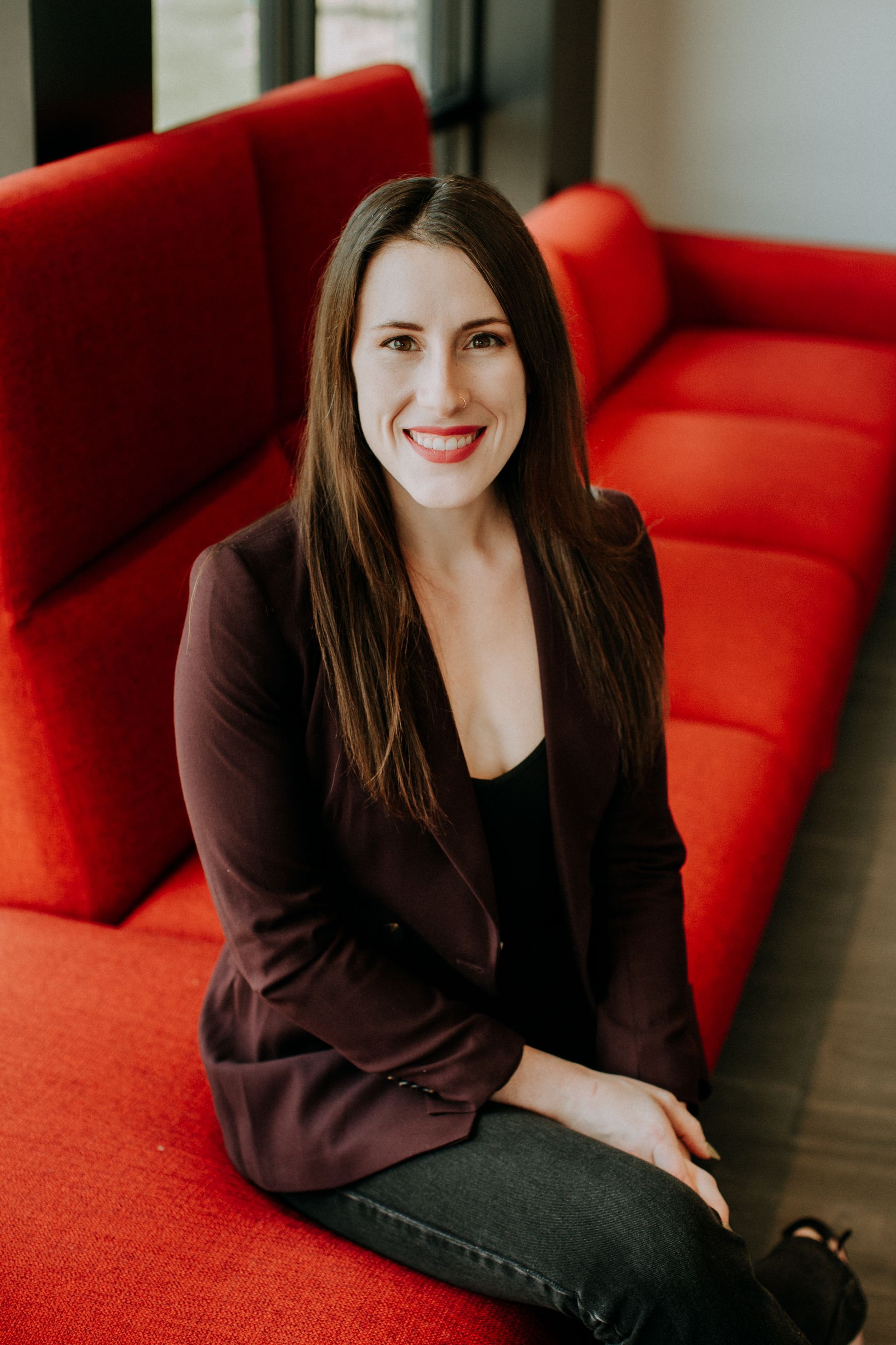 Woman in a burgundy blazer sits on a red sofa, smiling at the camera.