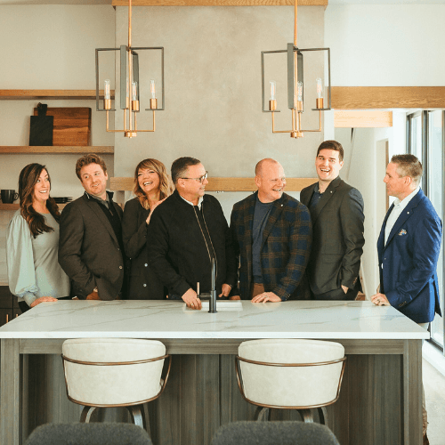 Group of seven people smiling in a modern kitchen. White countertop, gold light fixtures, and neutral tones.