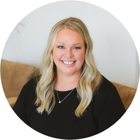Woman with blonde hair wearing a black top smiling; sitting on a brown couch.