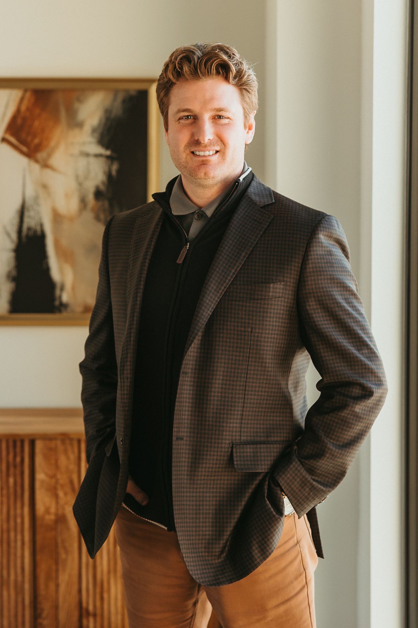 Man in patterned blazer, smiling, hands in pockets. Leaning against a window, art in the background.