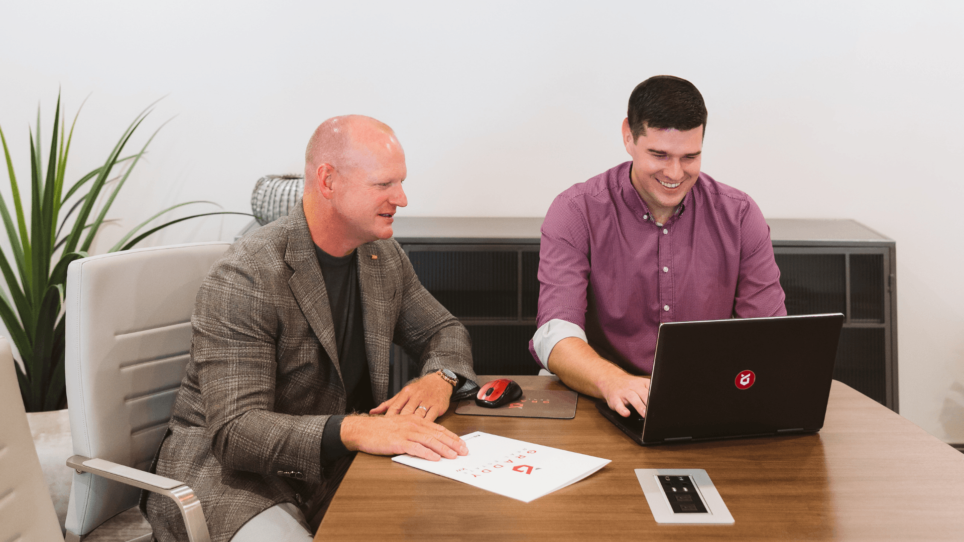 Two women in office setting reviewing documents, one points with pen, smiling.