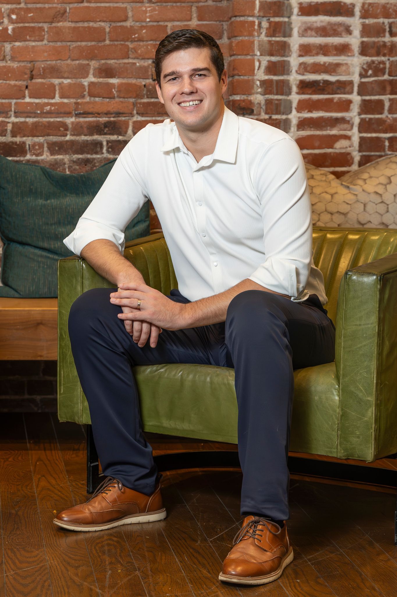 Man in white shirt and blue pants sits in a green chair. Brick wall background.