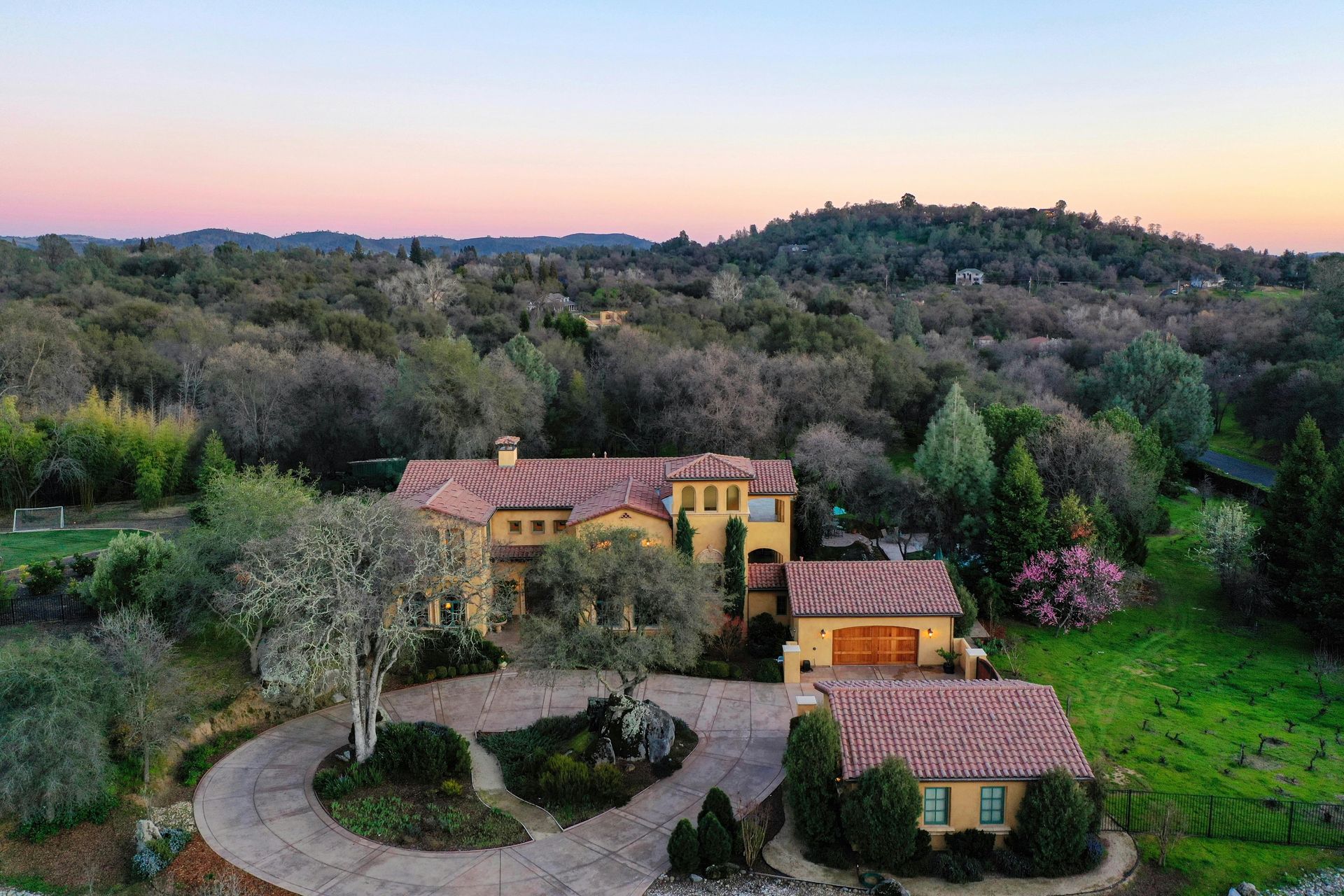 Yellow mansion with a terracotta roof, surrounded by trees and a circular driveway, sunset in the background.