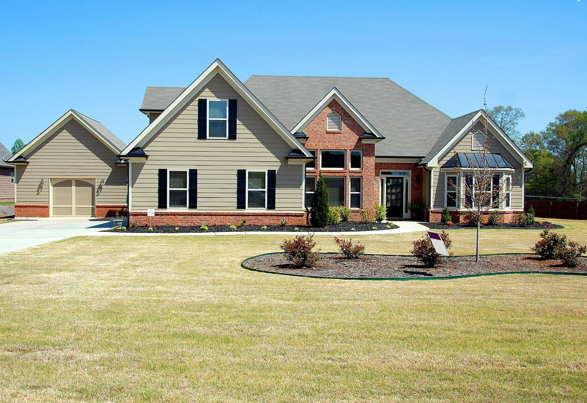 Tan house with brick accents, black shutters, and a two-car garage under a blue sky.