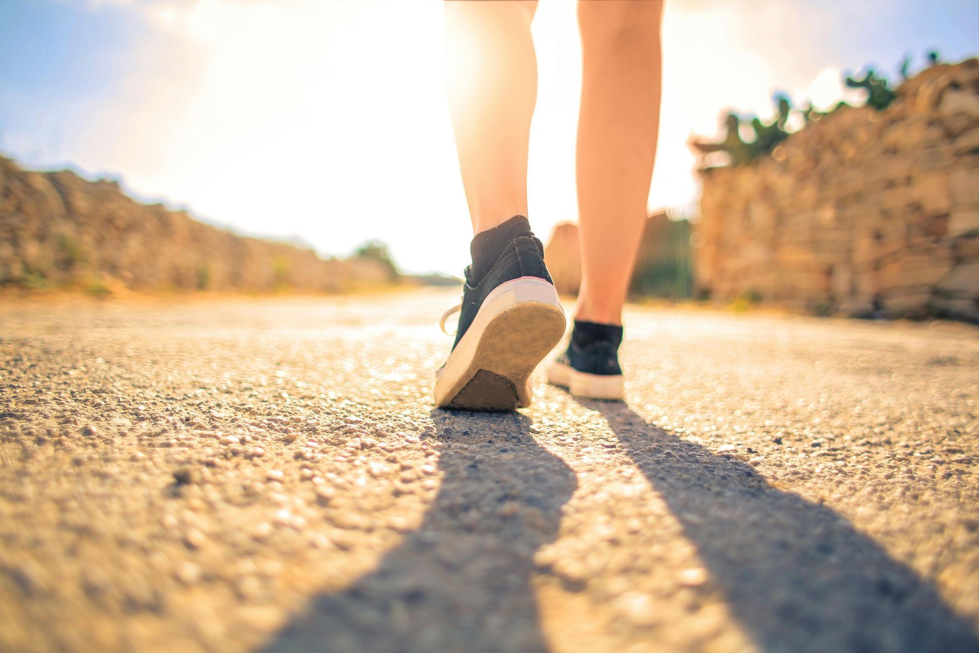 Person walking on a sunlit path, wearing black sneakers.