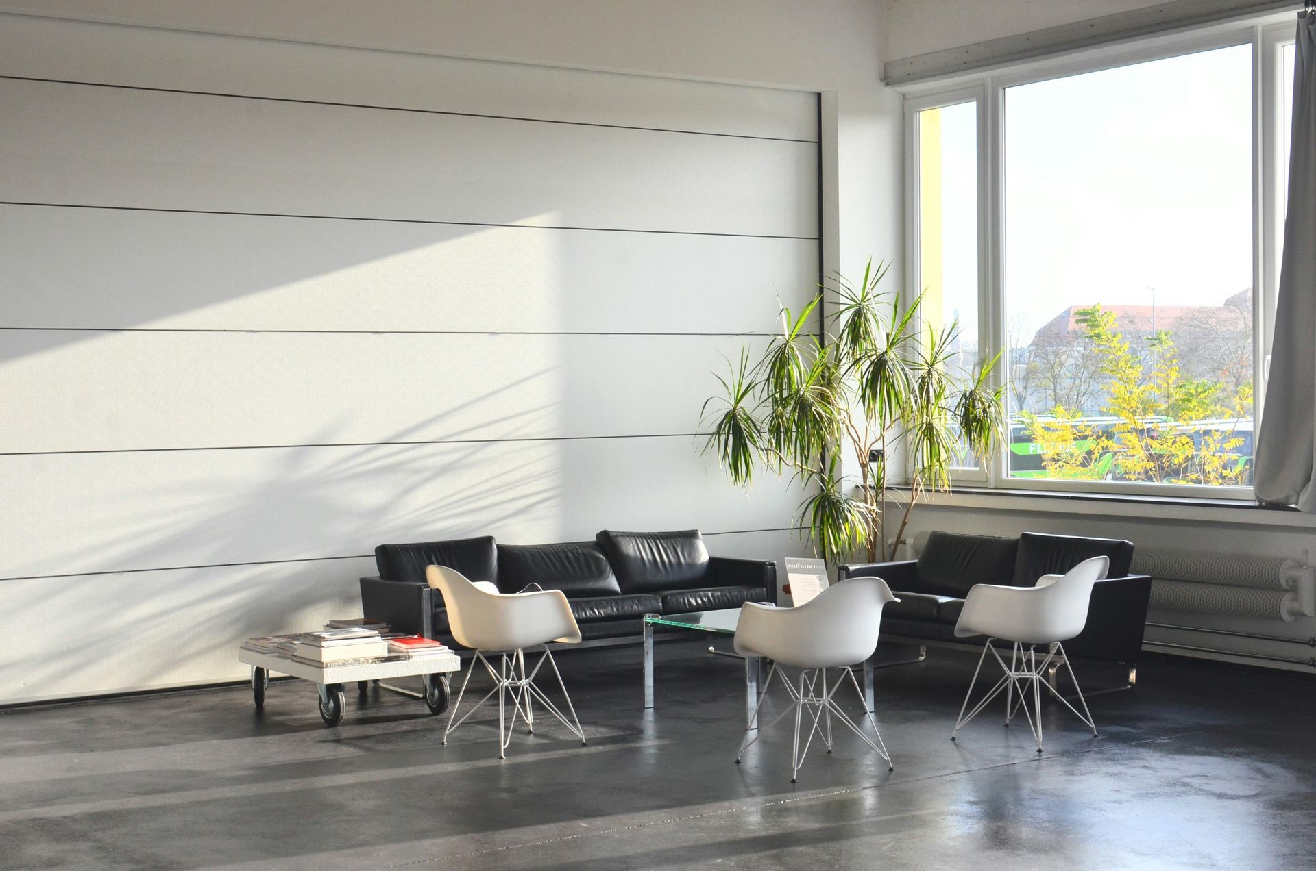 Lounge area with black couches, white chairs, and a glass table. Large window with sunlight and a potted plant.