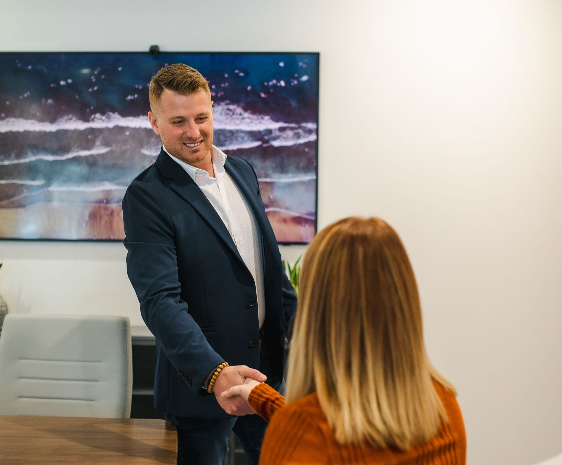 Man in a suit shakes hands with a person in an orange sweater at an office table. A large screen is visible.