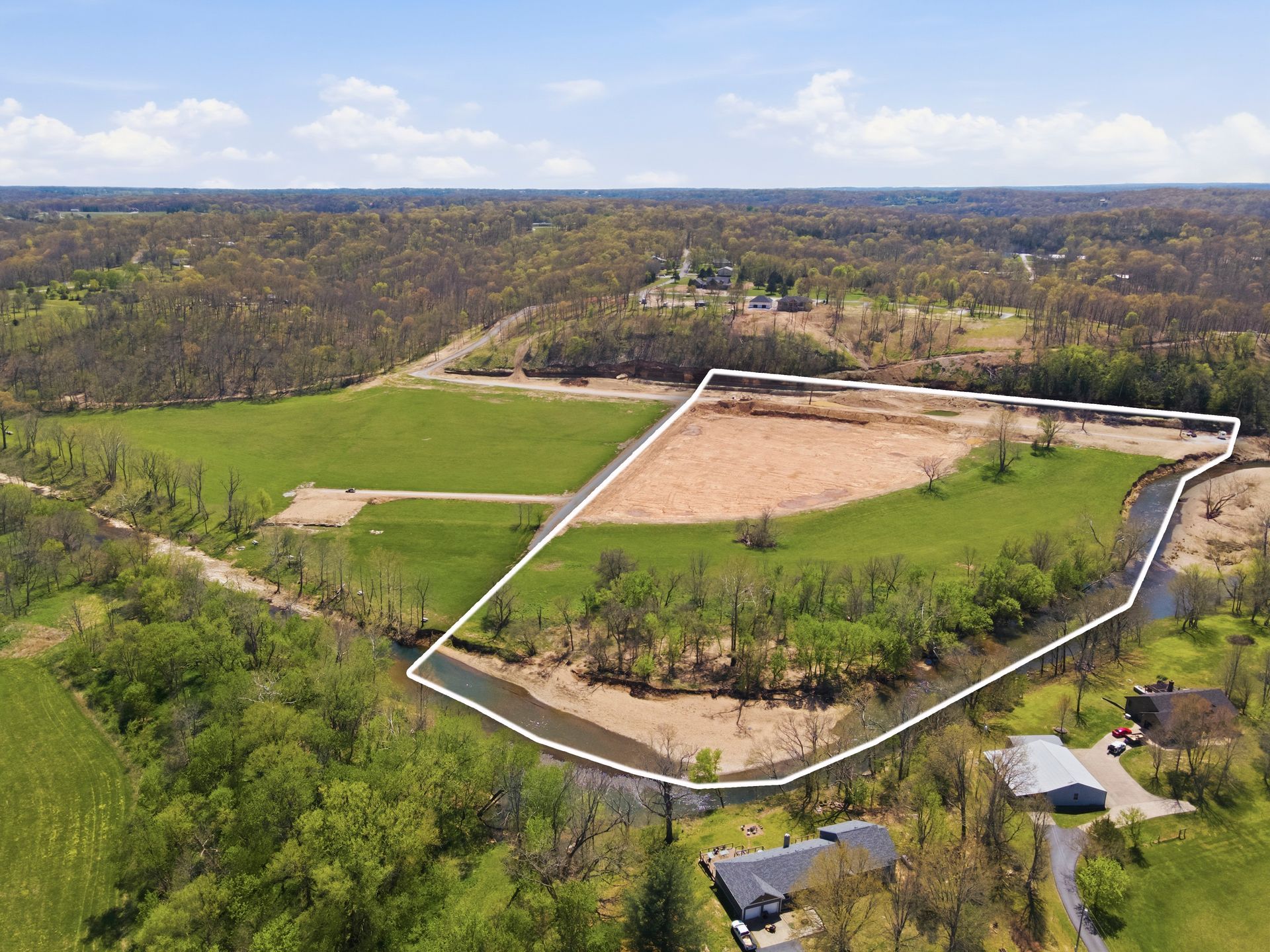 Aerial view of a fenced cleared field beside green woods and a few houses under a cloudy sky