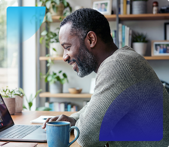 Man smiling while typing on a laptop at a desk with a coffee mug in a bright home office