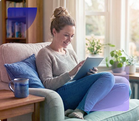 Woman smiling on a couch, using a tablet in a bright living room with a mug and plant nearby.