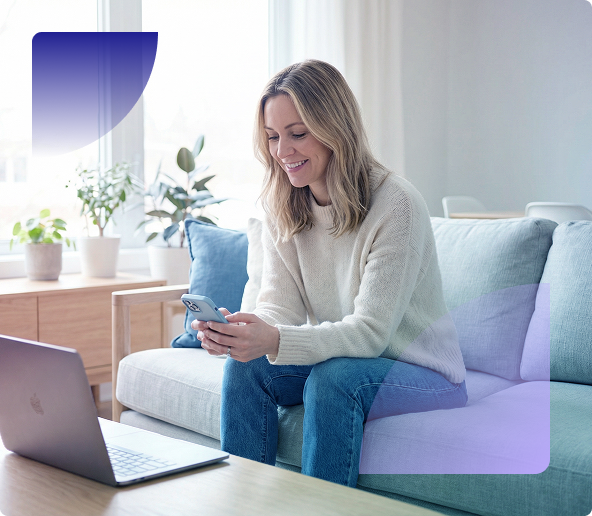 Woman sitting on a sofa using a smartphone beside a laptop in a bright living room