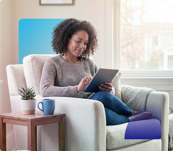 Woman seated in a beige armchair using a tablet beside a side table with a mug and plant in a bright living room