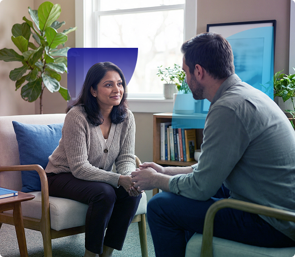 Two people talking on a sofa in a bright living room, holding hands during a counseling session.