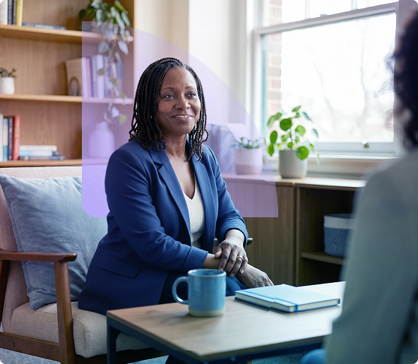 Woman seated in an office, smiling beside a blue mug and notebook near a bright window.