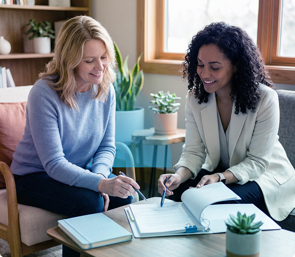 Two women taking notes together at a table in a bright living room with notebooks and a plant nearby