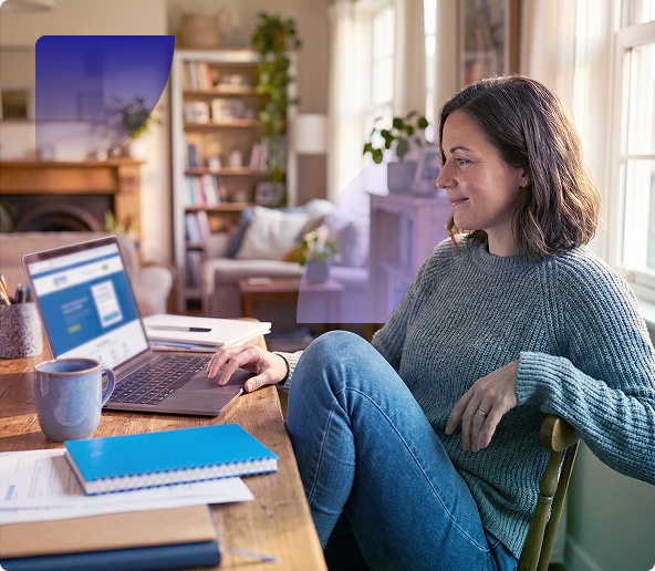 Woman working on a laptop at a cozy home desk with a mug, notebook, and living room in the background