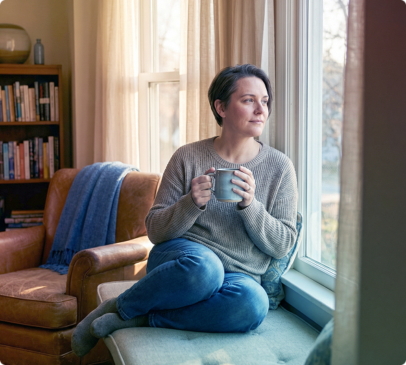 Person sitting on a window seat holding a mug, looking out beside a chair and bookshelf in a cozy room.