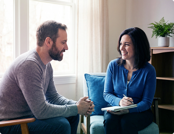 Two people talking and smiling in a bright living room, seated on chairs with a blue cushion between them