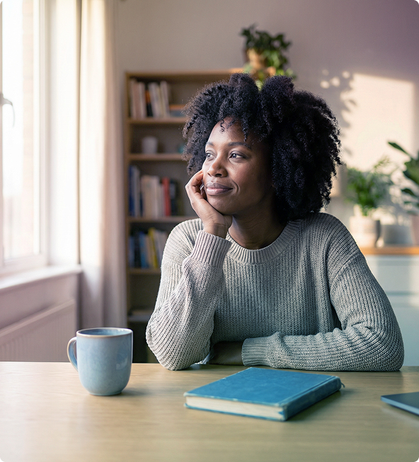 Woman seated at a table by a window, resting her chin on her hand beside a mug and notebook, smiling thoughtfully