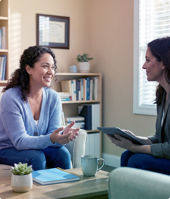 Two women talking in a cozy office, one holding a tablet and smiling across a coffee table.