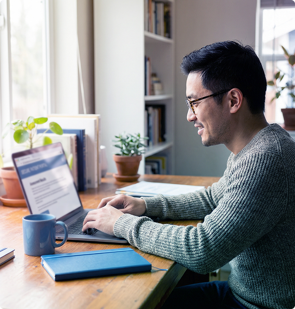 Man typing on a laptop at a desk by a window, with a mug, notebook, and potted plant nearby