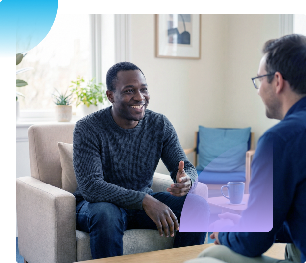Two people talking in a bright living room, one smiling on a sofa and the other seated across from him.