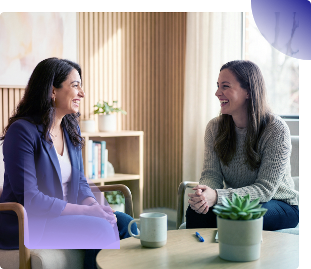 Two women chatting on a couch in a bright office lounge with coffee mugs and a small plant on the table