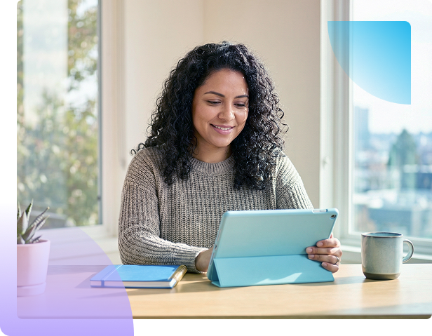 Person using a tablet at a desk with a mug and books, in a bright window-lit room.