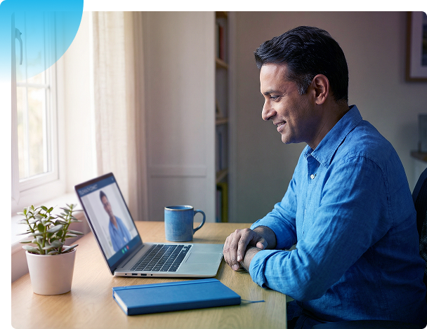 Man sitting at a desk using a laptop beside a blue notebook and coffee mug by a window