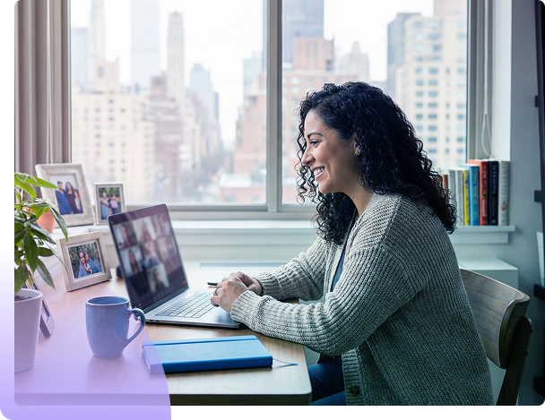 Woman working on a laptop at a desk by a bright office window with city buildings outside.