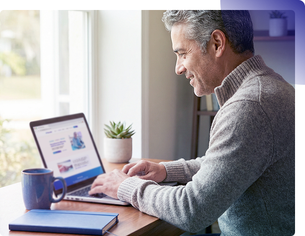 Man using a laptop at a desk by a window, with a coffee mug and small plant nearby