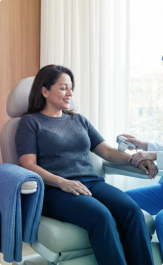 Woman sitting in a recliner, smiling as a caregiver checks her wrist in a bright room.