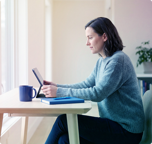 Woman in a blue sweater using a tablet at a table with a mug and notebook by a window