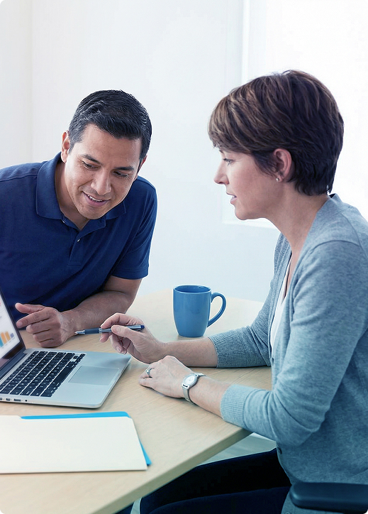 Two coworkers discussing work at a laptop with papers and a blue mug on a table