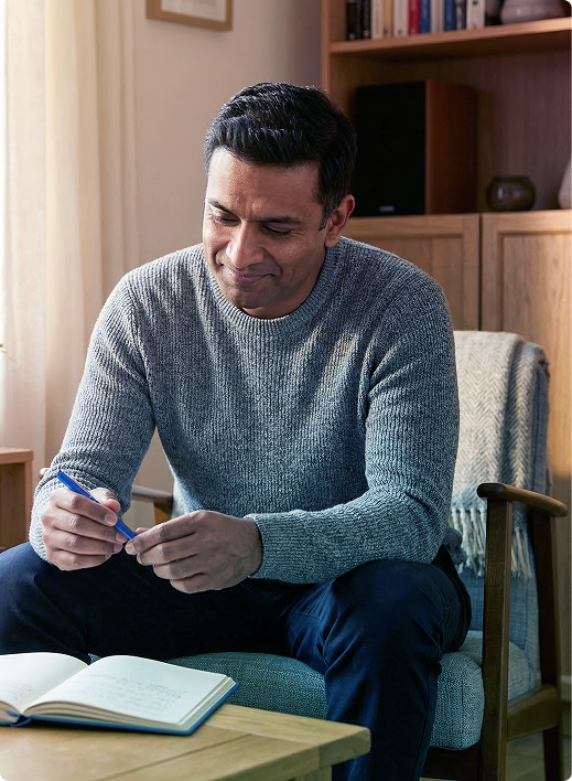 Man sitting in a chair, writing in a notebook with a pen in a cozy room.