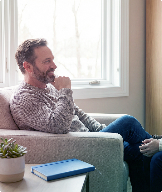 Man sitting on a sofa by a window, smiling in conversation with someone off-frame, with a notebook on the table.