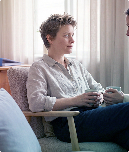 Two people sit facing each other in a bright room, talking quietly on chairs by a window.