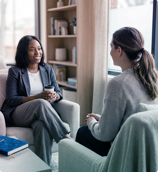 Two people in a bright office lounge talking across chairs, one holding a cup.